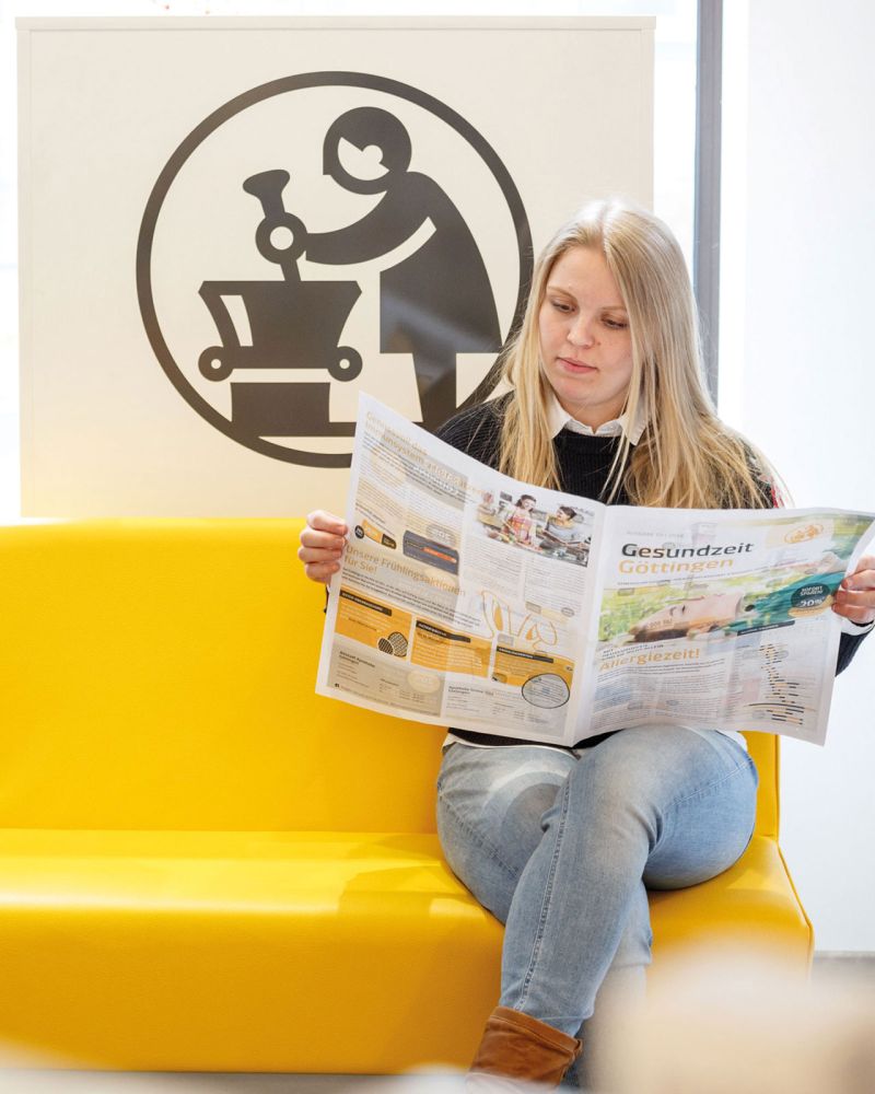 Lucht Apotheken Göttingen, Kundin sitzt mit Apothekenzeitung auf gelber Bank im Verkaufsraum der Apotheke, Logo im Hintergrund