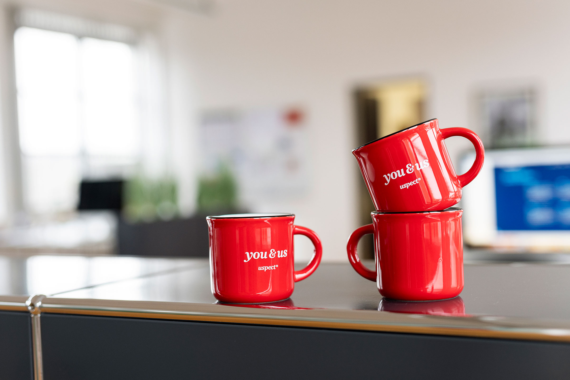 Rote Tassen mit dem Schriftzug "you & us" stehen auf einem Regal in der Agentur für Markenkommunikation