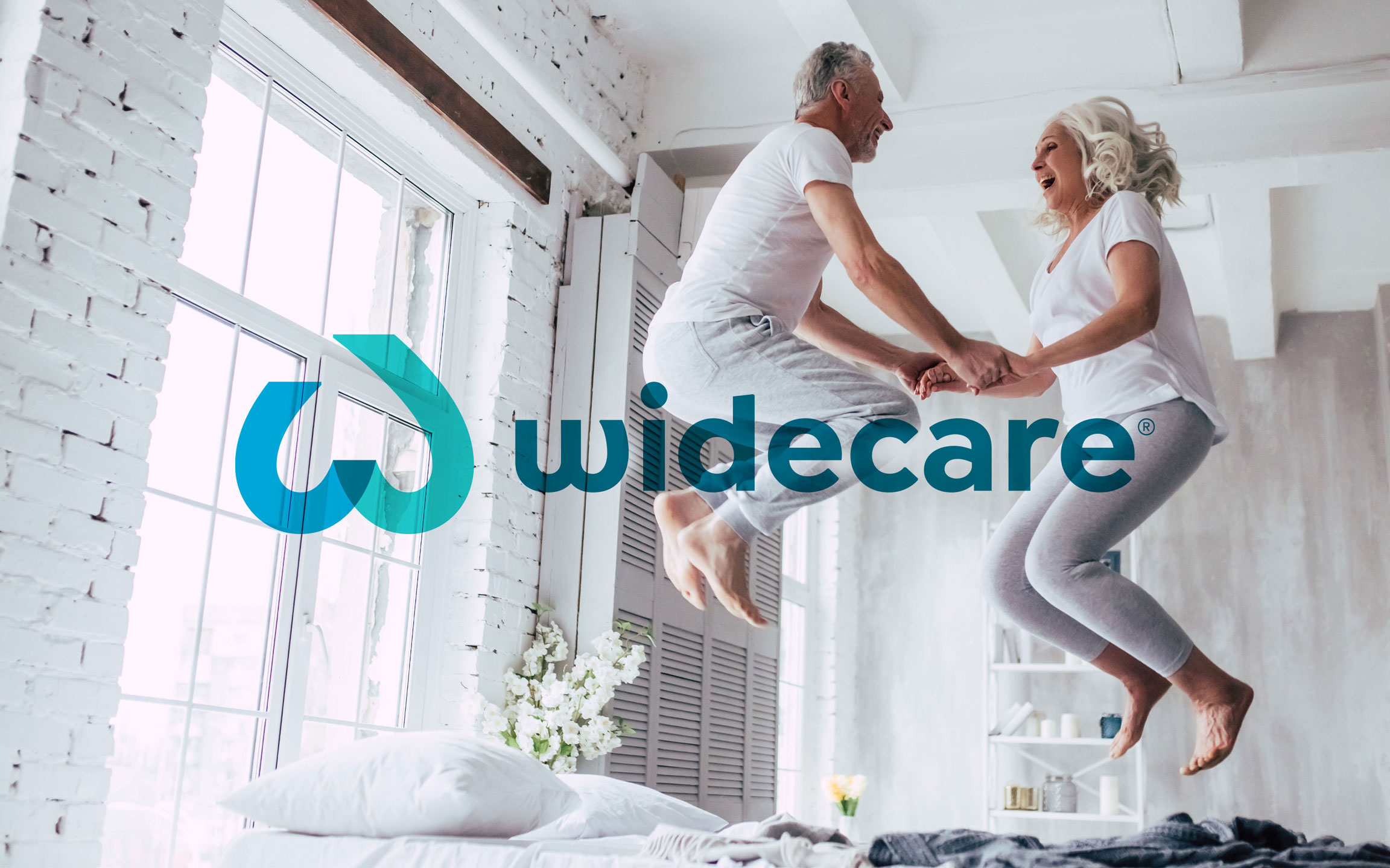 widecare Logo, Stuttgart, älteres Paar hüpft fröhlich auf Bett in Loft